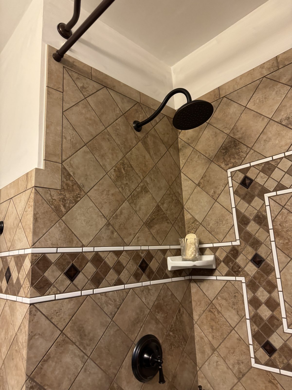Shower tile detail showing diagonal stone tile with dark accent diamond inlay border and oil-rubbed bronze controls