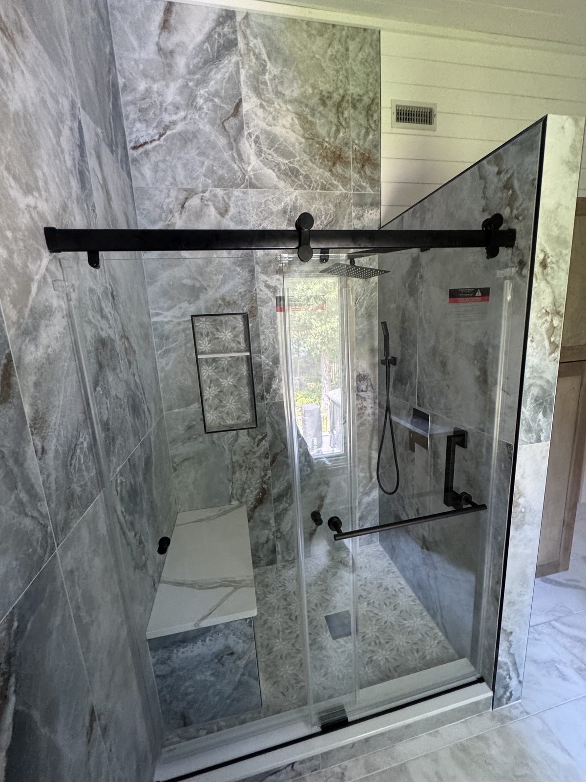 Glass shower enclosure with gray marble-look porcelain tile, matte black hardware, and natural light from window