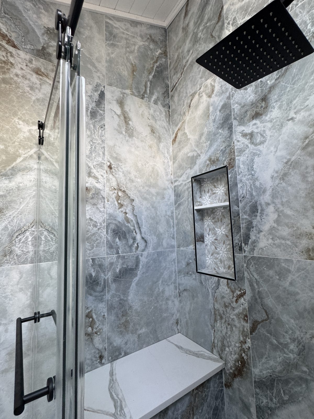 Walk-in shower with gray marble-look tile, matte black rain showerhead, recessed ceiling light fixture, and glass door