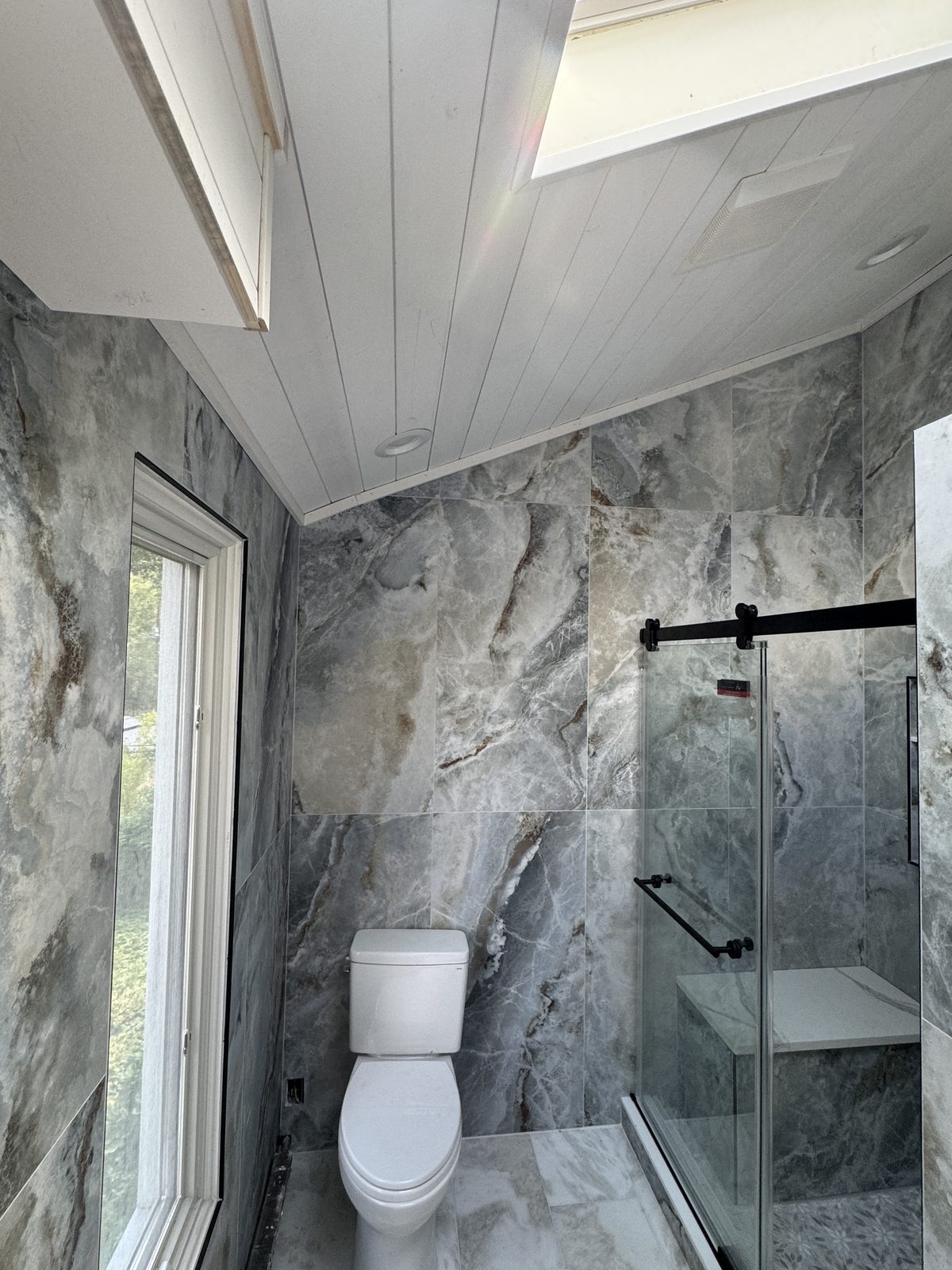 Full bathroom remodel with gray marble-look porcelain tile, glass shower enclosure, shiplap accent wall, and new window