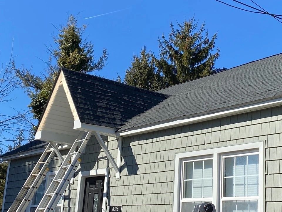 Newly installed dark shingle roof on gray home with ladder against gable